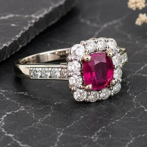 Könnte beinhalten: Nahaufnahme eines Weißgoldrings mit einem quadratisch geschliffenen, leuchtend roten Rubin als Mittelstein, umgeben von einem Halo aus kleineren, runden, klaren Diamanten. Das Band ist ebenfalls mit kleineren Diamanten besetzt. Der Ring wird auf einer dunklen, strukturierten Oberfläche präsentiert.