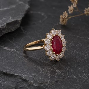 Könnte beinhalten: Ein goldener Ring mit einem großen, oval geschliffenen Rubin, umgeben von einem Kranz aus kleineren, klaren Edelsteinen. Der Ring hat ein schlichtes, poliertes Goldband. Die Schmuckstücke sind vor einem dunklen, strukturierten Hintergrund platziert.
