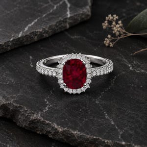 May include: A silver ring with a large oval-cut ruby center stone, surrounded by a halo of smaller diamonds. The ring's band is also encrusted with small diamonds, creating a sparkling effect. The ring is set against a dark, textured background.