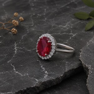 Könnte beinhalten: Ein silberner Ring mit einem großen, oval geschliffenen, tiefroten Edelstein, umgeben von einem Kranz aus kleineren, klaren Edelsteinen. Der Ring liegt auf einer dunklen, strukturierten Oberfläche, mit einem Zweig getrockneter Blumen und einem grünen Blatt im Hintergrund.