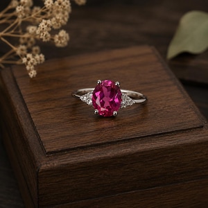 Könnte beinhalten: Ein silberner Ring mit einem großen, oval geschliffenen, leuchtend rosa Edelstein im Mittelpunkt. Der Ring wird von kleineren, dreieckigen Clustern weißer Edelsteine auf jeder Seite flankiert, auf dunkler Holzoberfläche. Schmuck.