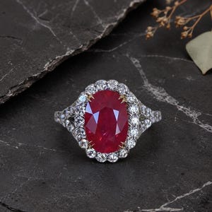 Könnte beinhalten: Ein silberner Ring mit einem oval geschliffenen, tiefroten Rubin, umgeben von einem Kranz kleiner Diamanten. Der Ring hat ein filigranes Design mit zusätzlichen Diamanten am Band. Präsentiert auf einem dunklen, strukturierten Untergrund.