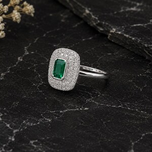 Könnte beinhalten: Ein rechteckiger Ring mit einem grünen Smaragd, umgeben von einem Kranz aus kleinen, klaren Edelsteinen. Der Ring ist auf einem silbernen Band gefasst. Der Ring wird auf einer dunklen, strukturierten Oberfläche präsentiert.