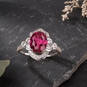 Könnte beinhalten: Ein silberner Ring mit einem großen, oval geschliffenen, rubinroten Edelstein als Mittelpunkt. Der Ring ist mit kleineren, klaren Edelsteinen und dekorativen Filigranarbeiten verziert. Der Ring ist vor einem dunklen, strukturierten Hintergrund platziert.