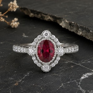 May include: A silver ring with a large oval ruby center stone, surrounded by a halo of smaller diamonds. The band is also set with small diamonds. The ring is displayed on a dark, textured surface.