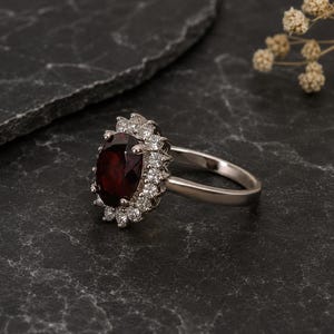 Könnte beinhalten: Ein silberner Ring mit einem großen, ovalen, tiefroten Edelstein, umgeben von kleineren, klaren Steinen. Der Ring liegt auf einer dunklen, strukturierten Oberfläche, mit getrockneten Blumen im Hintergrund. Der Ring hat ein dünnes, poliertes Band.