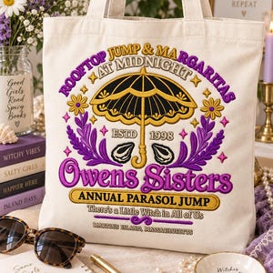 May include: A cream-colored canvas tote bag featuring purple and gold embroidery. The design includes a central parasol, the text "ROOFTOP JUMP & MARGARITAS AT MIDNIGHT", "OWENS SISTERS", "ANNUAL PARASOL JUMP", "There's a Little Witch in All of Us", and "MARTHA'S ISLAND, MASSACHUSETTS".
