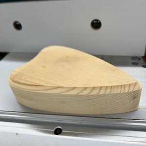 May include: A light brown wooden heart-shaped blank, with a slightly rounded edge, is sitting on a white surface.