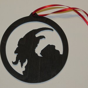 May include: Black wooden Christmas ornament with a silhouette of a goat-like creature with horns and a beard. The ornament has a red ribbon attached.