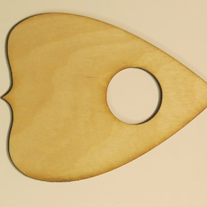May include: A wooden planchette in the shape of a heart with a circular cutout in the center. The planchette is light brown and has a smooth surface.