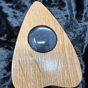 May include: A wooden Ouija board with a clear plastic window in the shape of a heart. The board is light brown with a wood grain pattern.