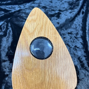 May include: A wooden Ouija board with a clear plastic window in the center. The board is shaped like a heart and has a dark brown stain.