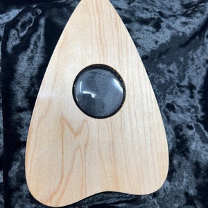 May include: A light-colored wooden planchette with a rounded triangle shape. It features a dark, circular viewing lens set in the center. The planchette is designed for use with a Ouija board.
