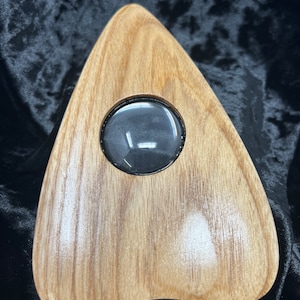 May include: A wooden triangular-shaped planchette with a round, clear glass window in the center. The planchette is a light brown color with a wood grain pattern.