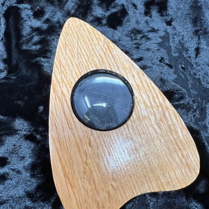 May include: A wooden Ouija board with a clear plastic window in the center. The board is shaped like a teardrop and has a light brown wood grain.