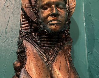 May include: A bronze-colored bust sculpture of a person with their eyes closed. The sculpture is decorated with metal accents and has a halo of nails around the head.