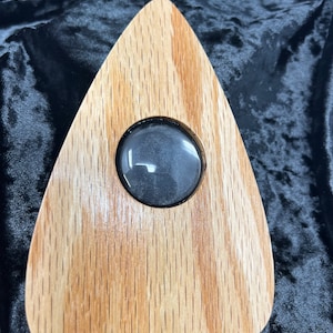 May include: A wooden planchette with a circular window, used for communicating with spirits. The planchette is shaped like a heart and has a light brown wood grain.
