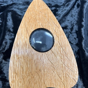 May include: A wooden triangular-shaped planchette with a circular glass window in the center. The planchette is made of light brown wood with a grain pattern.