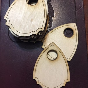 May include: A stack of three wooden planchette boards for use with a Ouija board. The planchette boards are shaped like a teardrop with a round hole in the center. The boards are light brown in color and have a smooth, polished finish.
