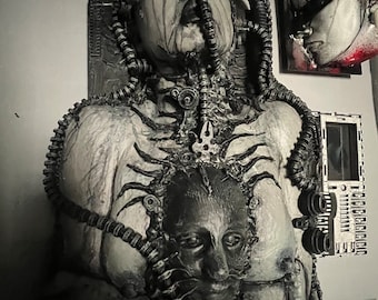 May include: A black and white sculpture of a human figure with a mechanical, alien-like appearance. The figure has a second face embedded in its chest, and is covered in tubes and wires.