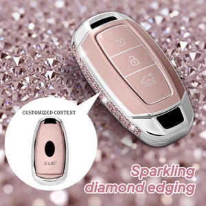 May include: A rose gold car key fob cover with a silver trim and sparkling diamond edging. The key fob has three buttons and the word "HOLD". The image also shows a smaller key fob with the text "CUSTOMIZED CONTENT".