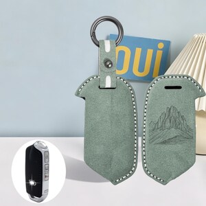 May include: A sage green suede key fob cover featuring white contrast stitching and a dark metal ring. One side displays a detailed mountain range line drawing. A small inset shows a black car key fob with silver buttons and a metallic base.