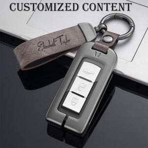 Puede incluir: Un llavero de coche gris plomo con tres botones blancos, sujeto a un llavero de cuero. El llavero está grabado con el nombre "Elizabeth Taylor" y el texto "CUSTOMIZED CONTENT" está en la parte superior.