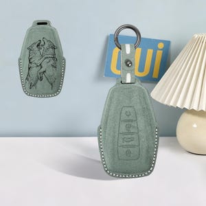 May include: A sage green suede car key fob cover featuring a detailed embossed graphic of two wolves howling and interacting. The cover includes a matching strap with a metal ring and a snap button, with white contrast stitching along the edges.