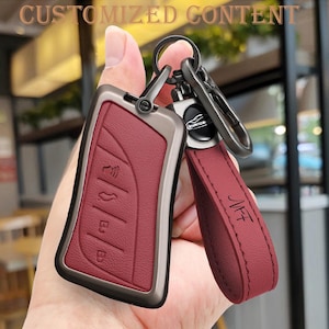 Puede incluir: Funda de llave de coche burdeos con marco de metal y correa de cuero a juego. El llavero tiene botones en relieve y un llavero de metal. La correa está grabada con las letras "NFT". Las palabras "CUSTOMIZED CONTENT" están en la parte superior.