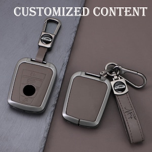May include: Two car key cases with keychains. The cases are gray with a silver-colored metal frame. One keychain has the text "CUSTOMIZED CONTENT" and the other has the initials "MFF" on the leather strap.