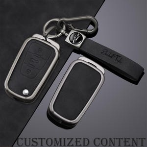 Könnte beinhalten: Ein Set schwarzer Leder-Schlüsselanhänger mit silberfarbenen Metallrahmen und -beschlägen. Einer der Anhänger hat ein Schlüsseldesign mit Knöpfen, ein anderer einen Lederriemen mit dem eingravierten Namen "Elizabeth Taylor". Der Text "CUSTOMIZED CONTENT" befindet sich unten.