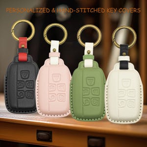 以下が含まれることがあります： ブロンズ色のリングが付いた4つのキーカバー。PERSONALIZED & HAND-STITCHED KEY COVERSの文字入り。カバーは、赤ストラップ付きの黒、白ストラップ付きの薄いピンク、白ストラップ付きのセージグリーン、黒ストラップ付きのクリーム色です。