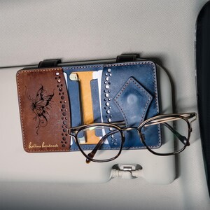 May include: A leather car visor organizer featuring a brown section with a butterfly engraving and the text hallmo handmade, alongside a blue section with card slots and a diamond-shaped tab holding a pair of brown-rimmed eyeglasses.