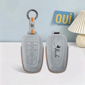 May include: Two light gray car key fob covers with orange stitching. One cover features a metal ring and orange strap, while the other displays the text Abcde with a small heart icon. The covers have a textured finish and molded button outlines.