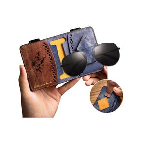 May include: A blue and brown leather glasses case featuring a butterfly engraving, perforated edges, and a yellow interior slot. The case holds black aviator sunglasses. A circular inset shows a brown leather tab with a yellow square snap closure.