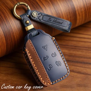 May include: A navy blue leather car key cover with brown accents and gold hardware. The cover has embossed symbols and the text "Be blessed" on the strap. The words "Custom car key cover" are at the bottom.