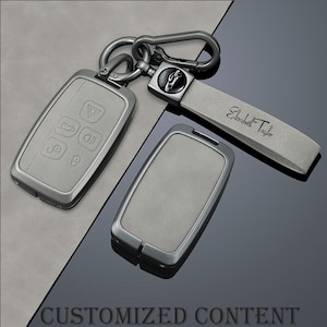 May include: A set of gray car key fobs with a matching key chain. The key fobs have a metallic frame and a soft, textured surface. The key chain includes a strap with the name "Elizabeth Taylor" and a car logo.