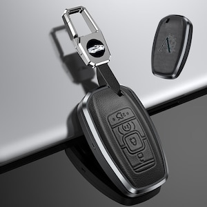 May include: A black leather car key fob with a silver metal trim and a silver key ring. The key fob has embossed buttons and a small car emblem on the key ring. A second, smaller black key fob with the letter "A" is also visible.