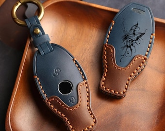 May include: Two oval-shaped leather key fob covers in navy blue and brown. One cover features a brass ring and a central circular cutout, while the other displays a black butterfly illustration. Both items are stitched with thick tan thread.