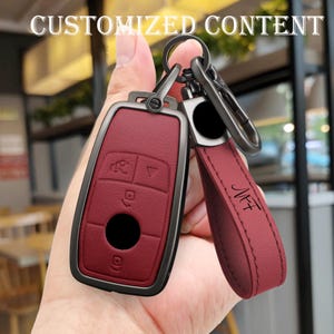 May include: A burgundy car key fob with a black metal frame and a matching leather strap. The key fob has several buttons and a circular black button. The strap has the initials "MFF" printed on it. The text "CUSTOMIZED CONTENT" is at the top.