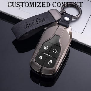 Peut inclure: Un porte-clés de voiture gris métallique avec une lanière en cuir noir. La lanière est gravée du nom "Mark Frederick". Le porte-clés comporte des boutons noirs avec des icônes blanches. Les mots "CUSTOMIZED CONTENT" sont en haut.