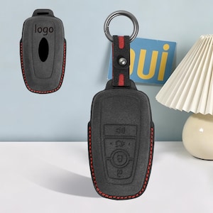 May include: A black key fob cover with red stitching and a matching strap featuring a metal ring. The cover has embossed button icons and a cutout for a logo. The design includes a red accent stripe on the strap, which is secured with a metal stud.