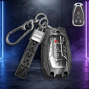 Puede incluir: Una funda protectora negra para llave de coche con acabado metálico, que incluye una correa de cuero negro a juego y un mosquetón de metal. La placa metálica en la correa tiene grabado el nombre Name. Se incluye una vista separada de la llave.