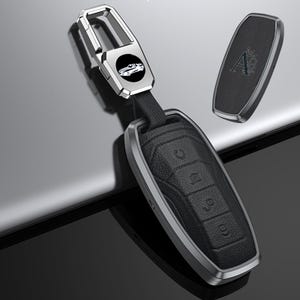 May include: Black leather car key fob with a silver metal frame and accents. The fob has buttons for various functions. A separate black leather key cover with a silver frame and a logo is also shown. A silver key chain with a car logo is attached.