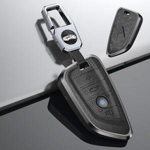 May include: A black leather car key fob with a silver metal trim and a silver key ring. The fob has a textured surface and a blue button. An additional key fob with the letter "A" is also visible.