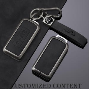 May include: Black leather key fob covers with a silver-tone metal frame. One key fob is attached to a key ring and a black leather strap with the name "Elizabeth Taylor" engraved. The text "CUSTOMIZED CONTENT" is at the bottom.