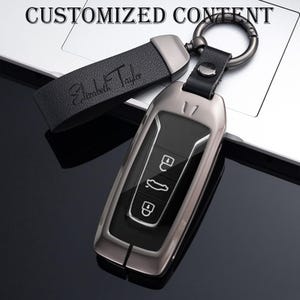 May include: A metallic car key fob with a black leather strap. The strap is embossed with the name "Elizabeth Taylor". The key fob has buttons for locking, unlocking, and other car functions. The words "CUSTOMIZED CONTENT" are at the top.
