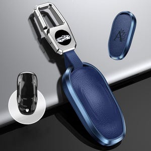 May include: A blue leather car key fob with a silver metal frame and a matching key ring. The fob has a car logo and a leather strap. An additional blue leather fob with the letter "A" is also shown.