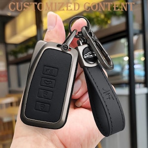 Peut inclure: Un porte-clés de voiture en cuir noir avec un cadre en métal argenté et un porte-clés assorti. Le porte-clés comporte quatre boutons et une lanière en cuir avec les initiales "MFT" en relief. Le texte "CUSTOMIZED CONTENT" est en haut.
