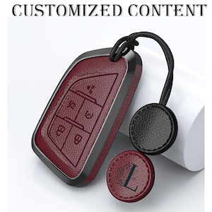 Puede incluir: Una funda de llavero de cuero burdeos con ribete negro y un cordón negro. La funda tiene símbolos de llave en relieve. También se incluye una etiqueta redonda a juego en burdeos y negro con la letra "L". El texto "CUSTOMIZED CONTENT" está en la parte superior.